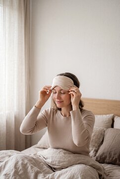 Beautiful woman waking up in a cozy bedroom, lifting a beige silk sleep mask and smiling after a good night's rest