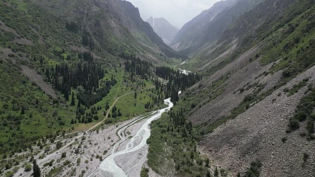 Aerial view of a winding river flowing through a deep mountain valley with steep rocky slopes, pine forests, and a dirt path in Bishkek, Bishkek City, Kyrgyzstan.