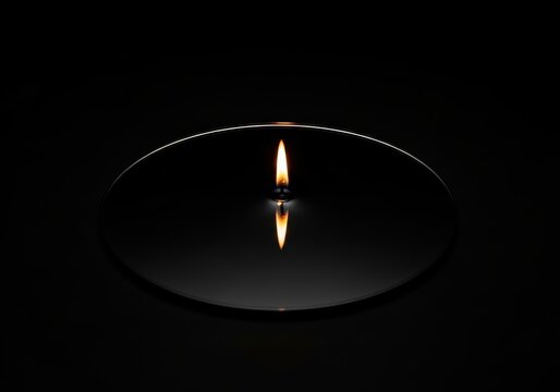 Burning candle with reflection on dark surface. Small golden flame glowing in deep black background. Symbol of memory, peace, hope, and meditation. Conceptual lighting design image.