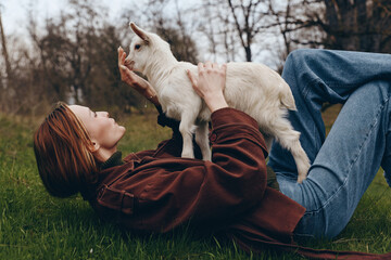 Obraz premium Young woman lying on grass plays joyfully with a white baby goat in an outdoor natural setting during a relaxed lifestyle moment.