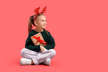 Naklejka premium Cute little girl in Christmas reindeer horns with gift book sitting on red background