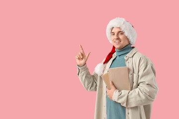 Naklejka premium Young man in Santa hat and with books pointing at something on pink background