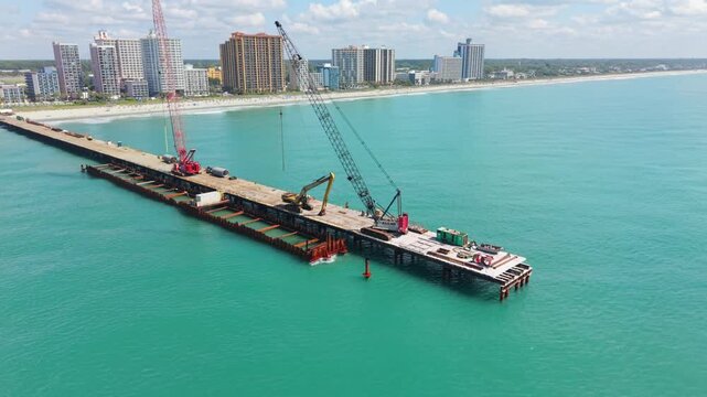 Construction cranes are actively lifting materials on a pier at Myrtle Beach, South Carolina, with the ocean and city skyline in view
