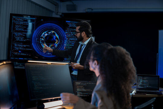 South Asian male and Black female software developers pair-program at dual monitors displaying network security algorithms at night.