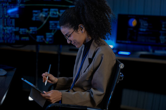 Black female data analyst cross-references code on a monitor with notes on a tablet in a blue-lit nighttime operations center.