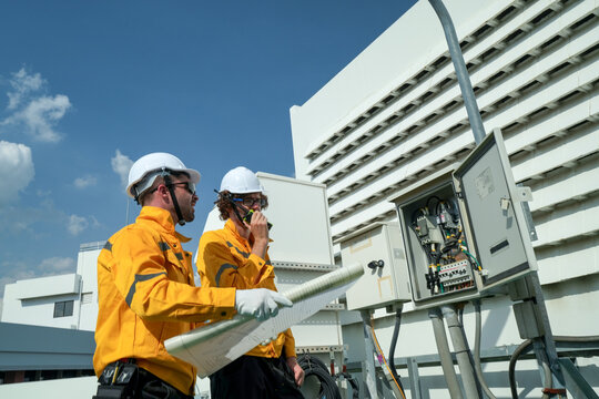 On-site view as a technician in an orange work jacket uses a laptop while servicing rooftop telecom and electrical equipment high above the urban skyline, capturing solar power generation.