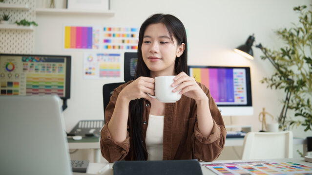Young graphic designer holding a coffee mug and reviewing printed design sheets in a bright creative workspace.