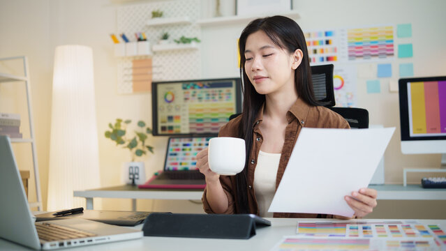 Young graphic designer holding a coffee mug and reviewing printed design sheets in a bright creative workspace.