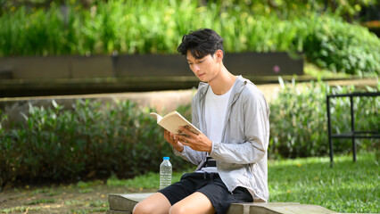 Obraz premium Young Asian Man Reading Book Outdoors in Green Park on Stone Bench