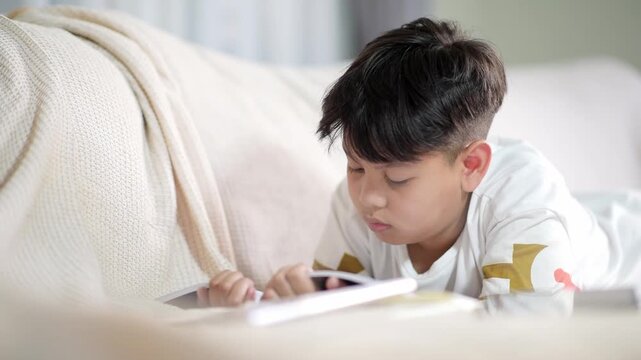 Focused Asian schoolboy reading physical book while resting on comfortable sofa. Concentrated preteen pupil studying traditional offline education materials and doing academic homework assignment