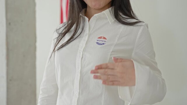 Woman in white shirt wearing voted sticker points finger to camera while presenting open palm, american flag visible on wall in building; pride civic duty.