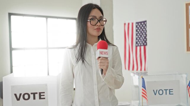 Woman reporter holding news microphone and speaking at polling station near vote booth and american flag; civic duty serious.