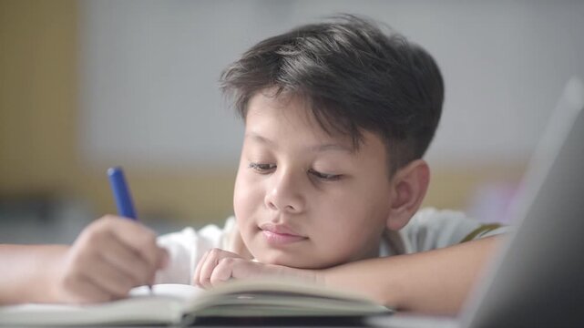 Smart Asian schoolboy writing notes thinking deeply before smiling brightly with new realization. Clever preteen pupil successfully solving difficult academic assignment