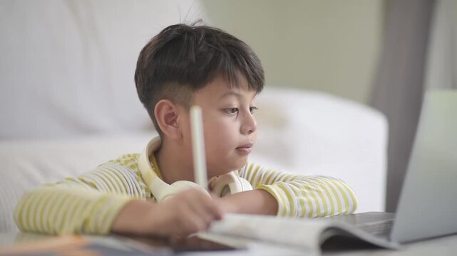 Smiling Asian schoolboy writing in workbook and waving hand at laptop screen. Cheerful gen alpha pupil communicating during interactive virtual online distance learning