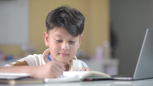 Focused Asian schoolboy with headphones around neck writing notes in workbook and smiling. Concentrated preteen pupil successfully finishing academic homework for modern edtech online course