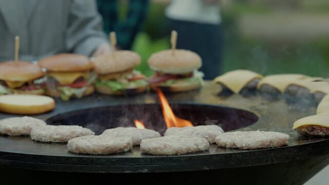Burger patties and cheeseburgers grilling over open flames on outdoor flat top cook surface