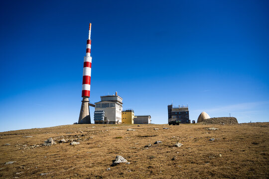 View of Botev Peak with its iconic red and white radio tower, mountain station buildings, and rocky terrain under a clear blue sky in Balkan Mountains, Lovech, Bulgaria.