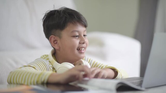 Cheerful Asian schoolboy enjoying interactive online learning on a laptop at home. The excited preteen waves and points at the screen, perfect for modern e-learning commercials