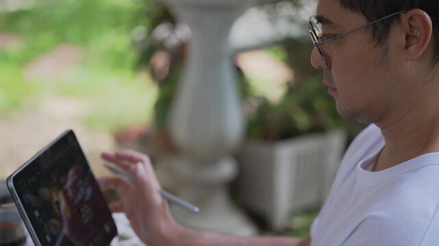 Side view of camera moving towards tablet as focused Asian man works in sunny garden in morning. Remote freelance workflow footage for tech and business media.
