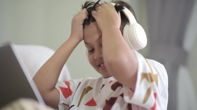 Frustrated Asian schoolboy grabbing his hair while struggling with difficult online homework. The stressed preteen wears headphones looking at a laptop, ideal for education themes