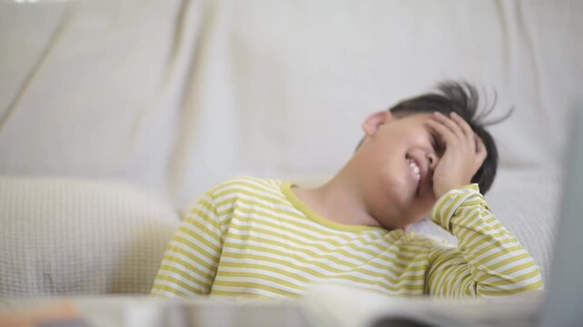 Tired Asian schoolboy resting chin on hand feeling overwhelmed during interactive online education course. Exhausted preteen pupil needing a short break from intense digital learning