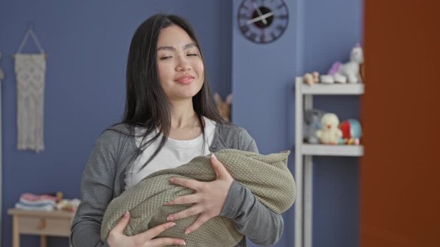 Woman cradling a swaddled baby, hands supporting the bundle in a nursery with toys and clock; bonding tenderness.