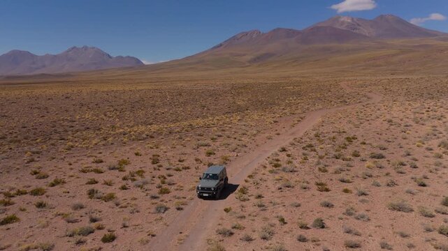 Compact offroad capable suv driving across dirt road desert track with mountains in backdrop, drone orbit follow