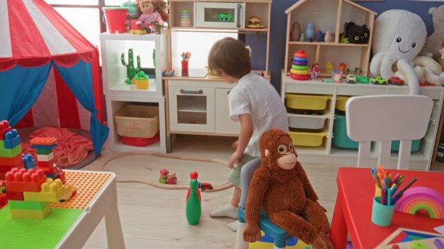 Boy picking up a plastic bowling pin and arranging toys beside a stuffed monkey in a colorful preschool playroom building; playful curiosity.