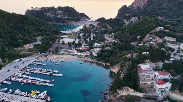Aerial view of Alipa marina port in Paleokastritsa, Corfu Island, Greece. Drone shot of moored boats, crystal clear turquoise sea water, lush green hills and coastal village roads during sunrise.