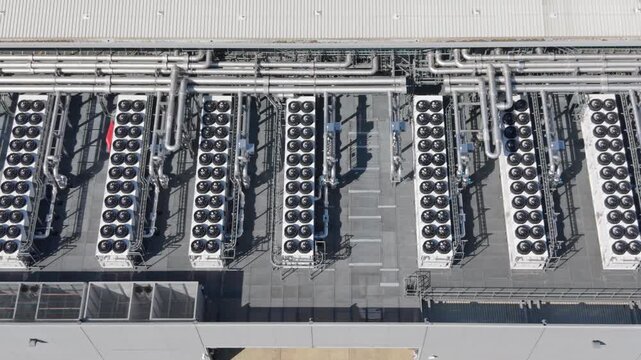 Overhead view of a data center facility rooftop. Numerous industrial cooling units, arranged in rows, with complex pipework, keep the servers cool on a sunny day.
