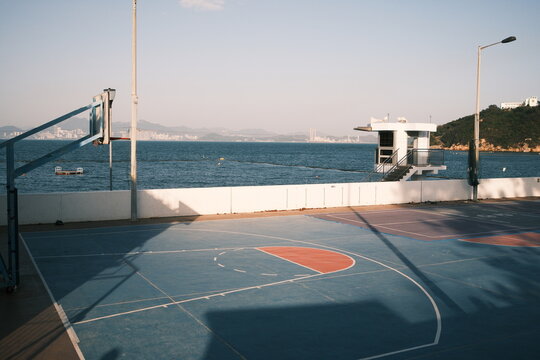 View of an outdoor basketball court with blue and red markings situated by the sea with a lifeguard tower and distant hills in Hong Kong, Hong Kong.