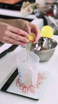 Vertical video. Food preparation. Baking ingredients. Female confectioner hand cracking raw egg in measuring cup on kitchen scale.