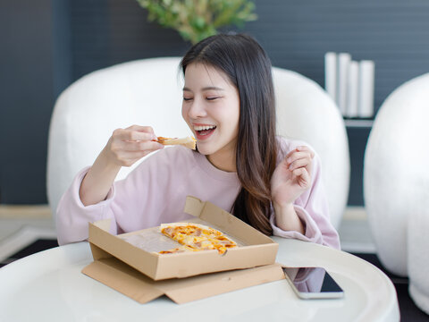 Asian beautiful cheerful young woman smiling eating delicious slice hot pizza delivery sitting in living room at home attractive girl feel happy relax enjoy weekend takeaway dinner takeout fast food.