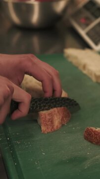 Vertical video. Cooking process. Cutting bread. Chef hands preparing ingredient for toasts on kitchen board. Worker slicing crunchy crust with knife.