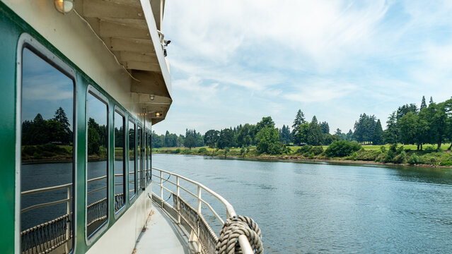 View of the Willamette River from the deck of a riverboat, featuring lush green trees along the riverbank under a bright blue sky in Portland, Oregon, United States.