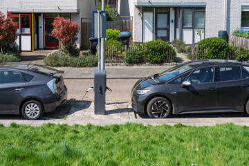 Obraz premium Two modern electric vehicles parked on a quiet residential street in the Netherlands, connected to a public charging station pillar. Sustainable urban transport infrastructure.