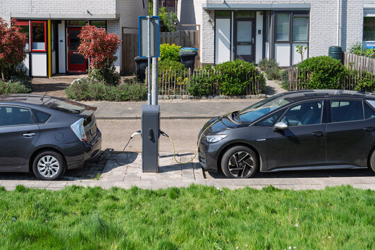 Two modern electric vehicles parked on a quiet residential street in the Netherlands, connected to a public charging station pillar. Sustainable urban transport infrastructure.