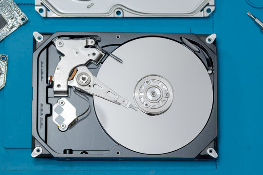 Disassembled computer hard drive inside view. Top view of an opened 3.5-inch desktop HDD showing internal components, shiny magnetic platter, actuator arm, and read-write head on a blue service mat