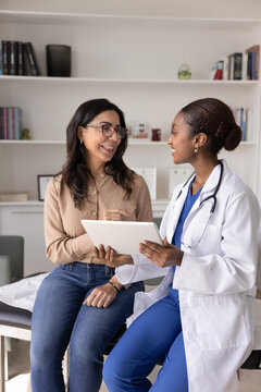 Positive young African doctor and senior Caucasian woman discussing good healthcare news for patient, sitting on medical couch, talking, Physician using digital gadget for consultation. Vertical shot