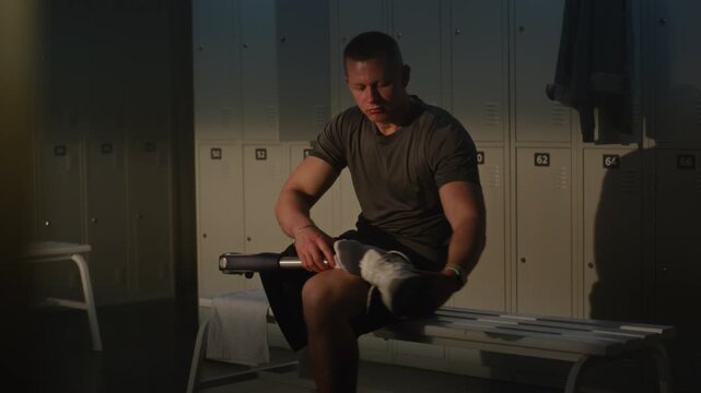 Young Man With Leg Prosthesis Sits on Locker Room Bench, Carefully Putting White Sneaker on Prosthetic Limb as Part of Pre Workout Routine in Sunlit Sports Facility. Resilience and Inclusion in Sport.