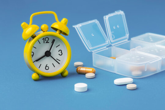 Yellow alarm clock with scattered pills and pill box on blue background, time to take medicine concept
