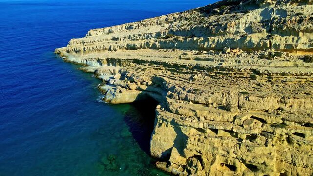 Stepped limestone cliffs rise above a wide blue sea with a dark coastal cave opening carved into the rock.