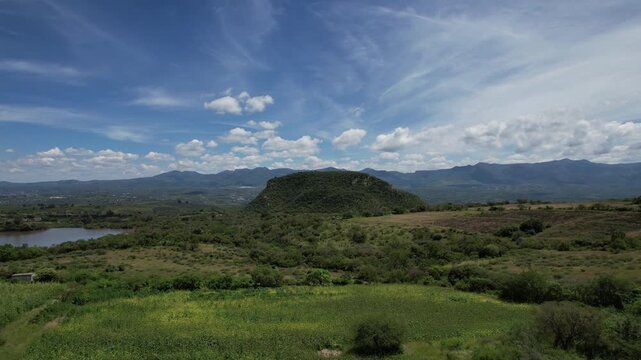 Aerial drone footage pulling back to reveal a scenic green landscape with a prominent flat-topped hill surrounded by fields, vegetation and distant mountain ranges beneath a vibrant blue sky filled wi