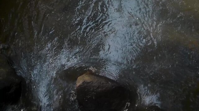Rushing river water cascades over dark rocks and stones in a mountain stream, demonstrating natural erosion patterns and scenic river flow within the Indonesian island landscape