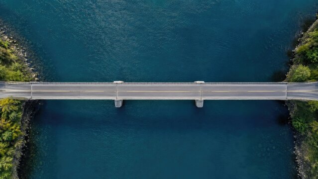 Aerial view of a straight two-lane bridge crossing deep blue water between green, tree-lined riverbanks