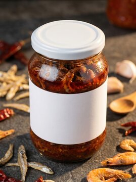 Blank label glass jar featuring savory dried shrimp and anchovy chili paste, surrounded by scattered ebi, ikan teri, garlic cloves, and a wooden spoon on a dark rough stone texture.