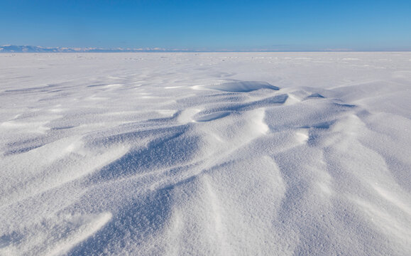 View of the frozen surface of Lake Baikal with snow ripples and distant mountains under a clear blue sky in Davsha, Buryatia, Russia.