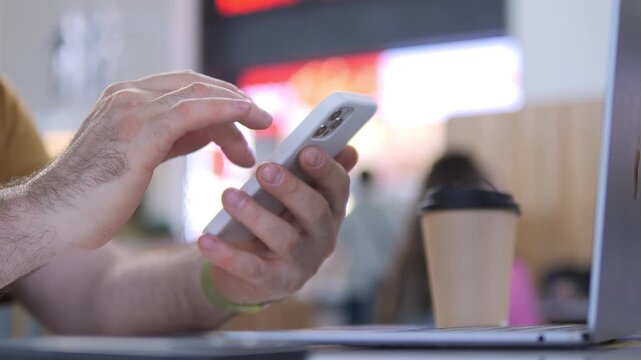 Close-up of hands scrolling smartphone while laptop sits nearby. Person focused, relaxed, multitasking in casual setting. Coffee cup beside, background shows cafe with soft lighting