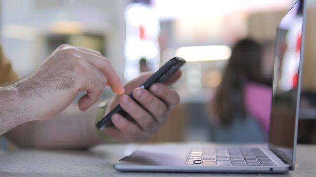 Hand uses smartphone while laptop sits nearby. Focus on digital multitasking in modern workspace. Background blurred to emphasize device interaction. Suitable for tech, business