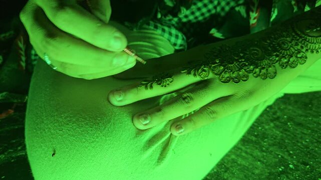 Close-up footage of a mehndi henna artist creating an intricate floral design on a woman's hand under decorative green LED lighting. Mehndi is an essential tradition during Indian weddings, festivals